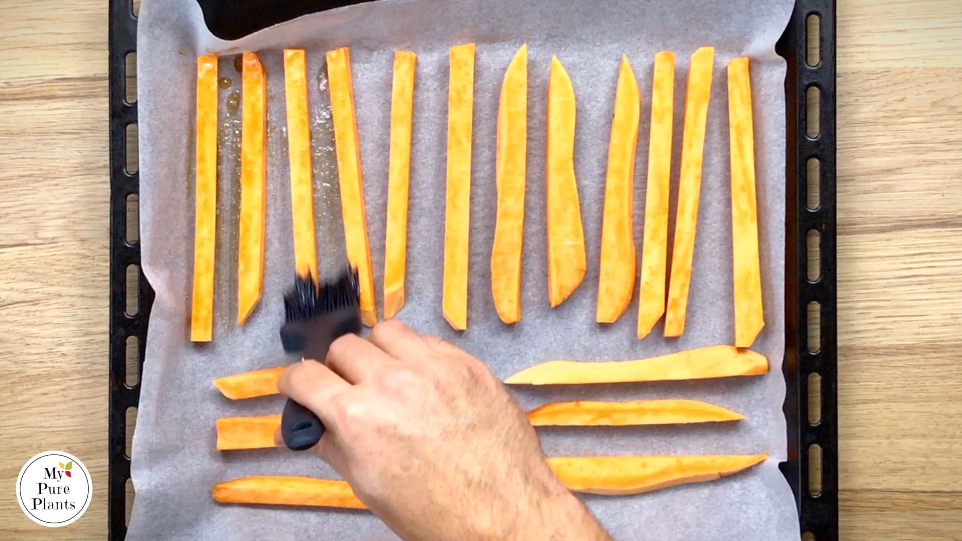 Sweet potato sticks are arranged in one layer on a parchment paper. A hand is brushing them with a marinade.