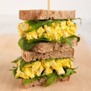A stack of two sandwiches. Both with brown sliced bread, arugula, and tofu cubes in thick yellow sauce to mimic egg salad.