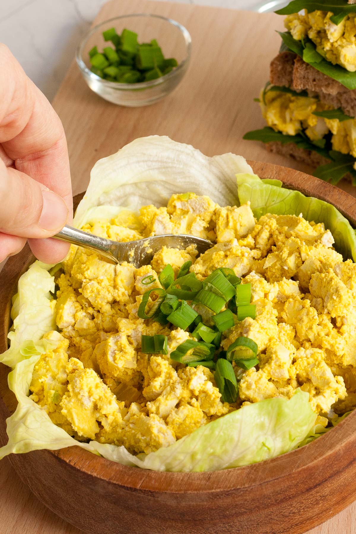 A wooden bowl with lettuce leaves topped with yellow egg salad. A hand is holding a spoon to take some.