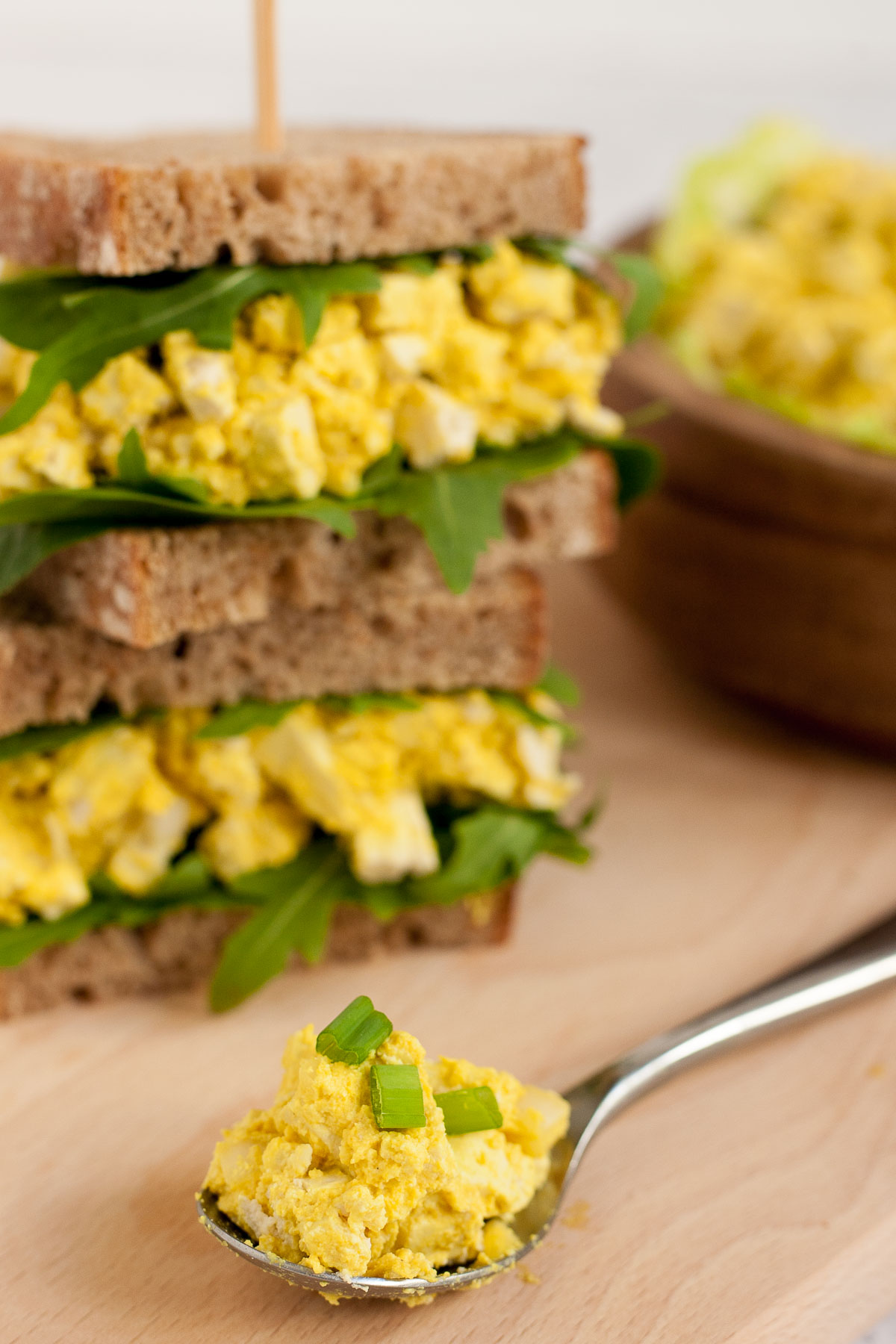 A spoon of yellow egg salad with a stack of two sandwiches. Both with brown sliced bread, arugula, and tofu cubes in thick yellow sauce to mimic egg salad.