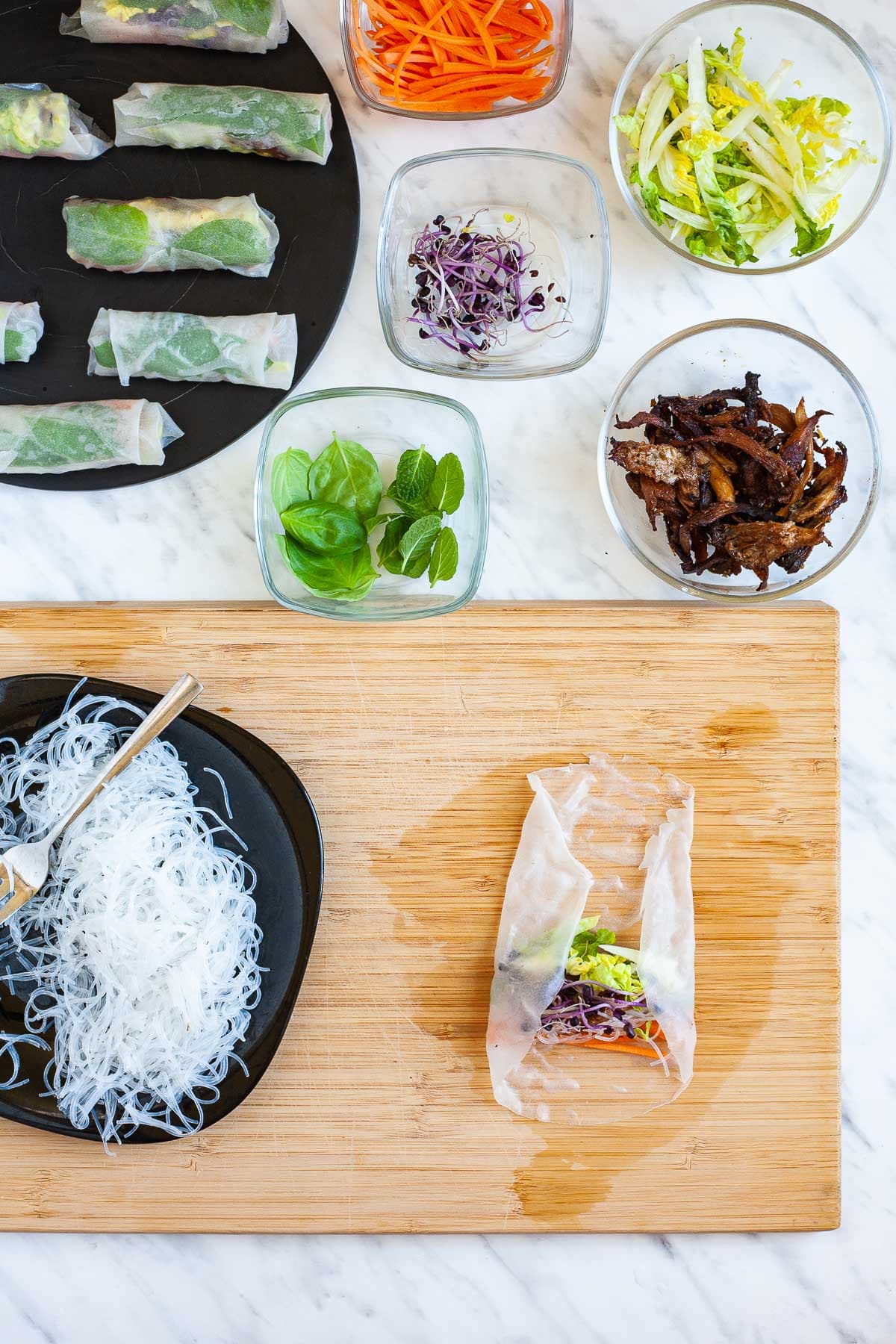 A wooden board with an opaque rice paper wrapper with filling, its side is folded to the middle. Fillings are around it in small glass bowls. Glass noodles on a black plate. The finished summer rolls are in the corner.