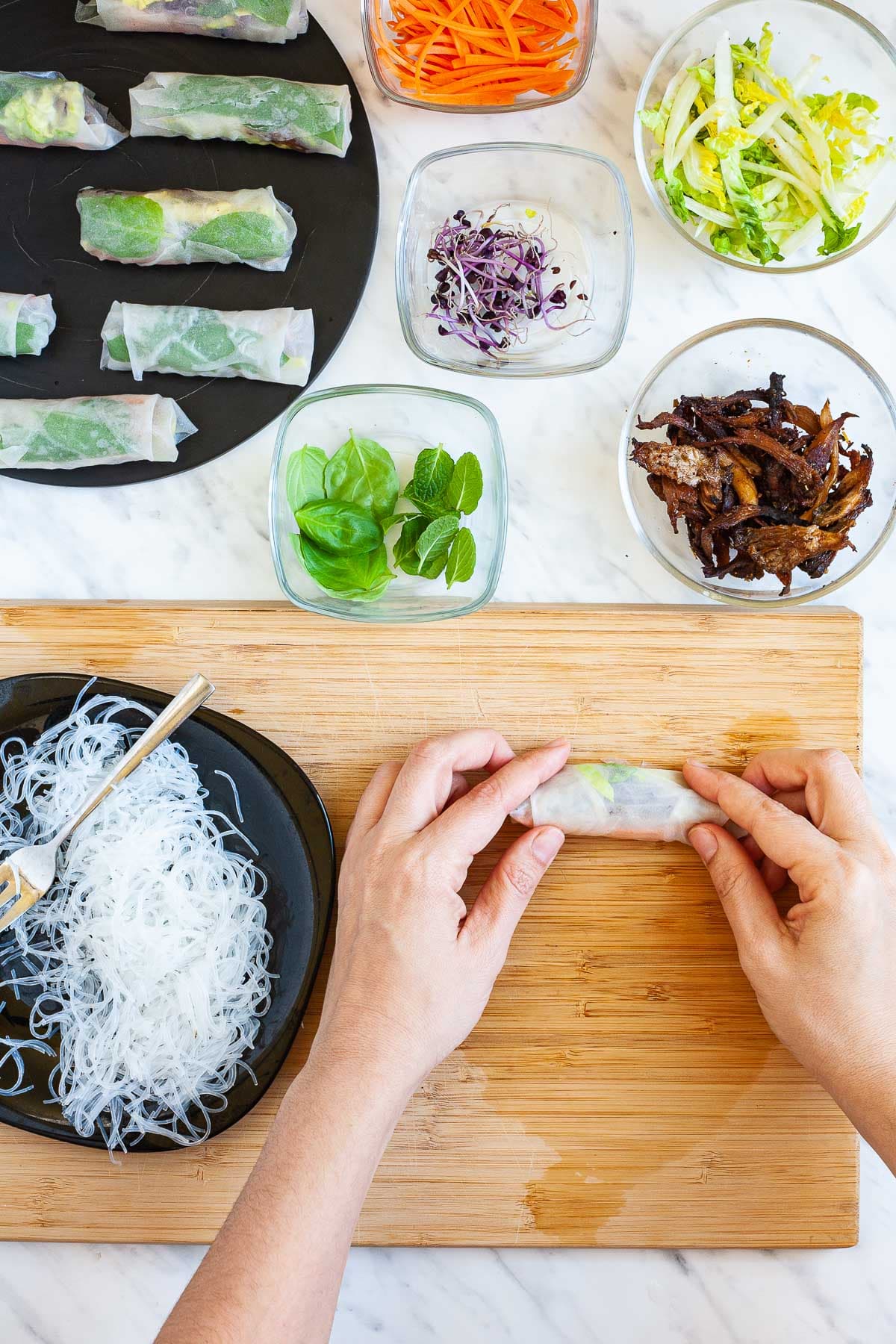 A wooden board with an opaque rice paper wrapper. A hand is rolling it from bottom to top. Fillings are around it in small glass bowls. Glass noodles on a black plate. The finished summer rolls are in the corner.