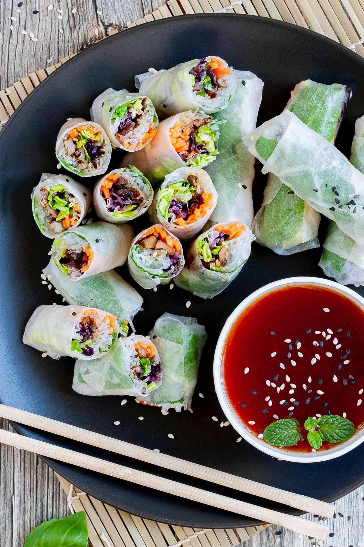 Opaque rice paper rolls filled with carrots, purple sprouts, thin glass. noodles and brown mushrooms are served on a large black plate. Several rolls are cut half so you can see the filling. Red sauce in a small white bowl is served next to it. 