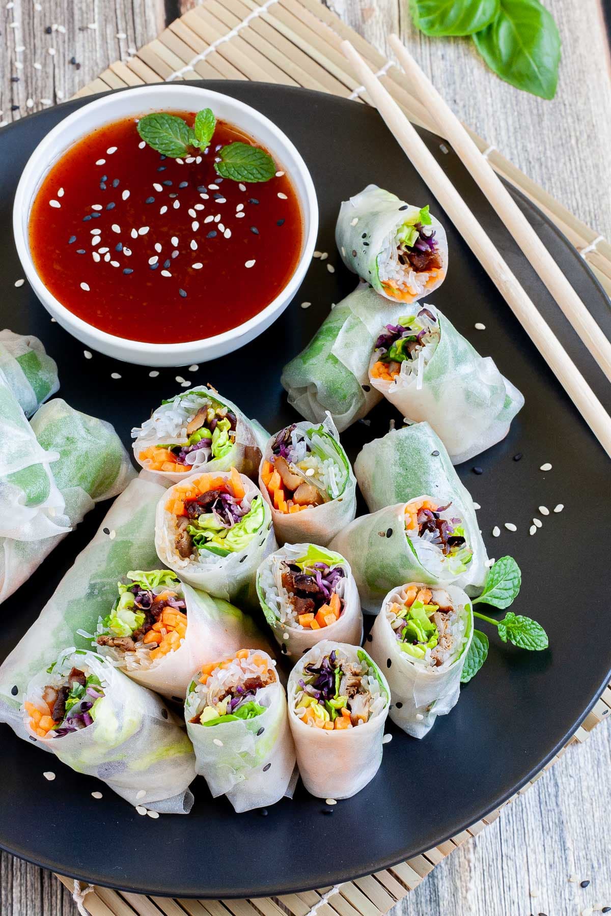 Opaque rice paper rolls filled with carrots, purple sprouts, thin glass. noodles and brown mushrooms are served on a large black plate. Several rolls are cut half so you can see the filling. Red sauce in a small white bowl is served next to it.