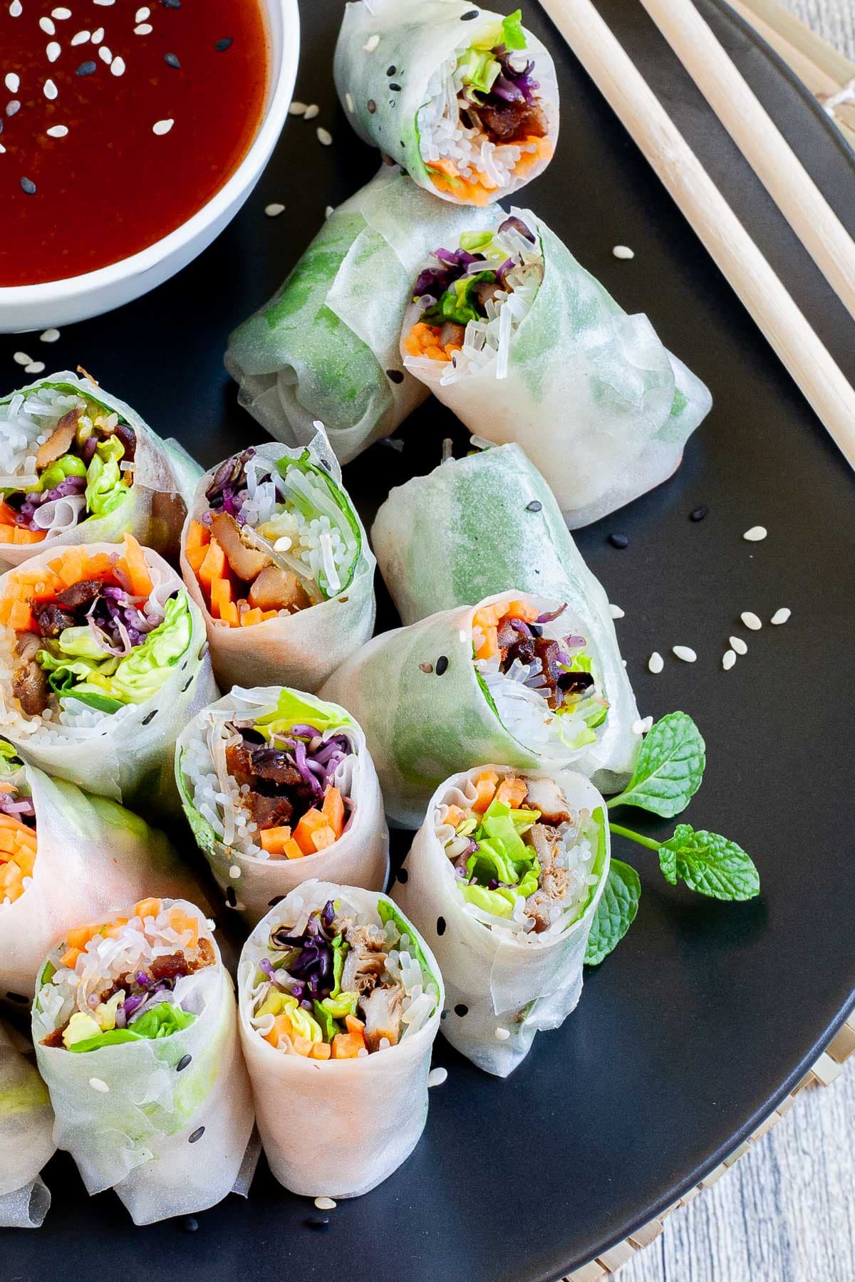 Opaque rice paper rolls filled with carrots, purple sprouts, thin glass. noodles and brown mushrooms are served on a large black plate. Several rolls are cut half so you can see the filling. Red sauce in a small white bowl is served next to it.
