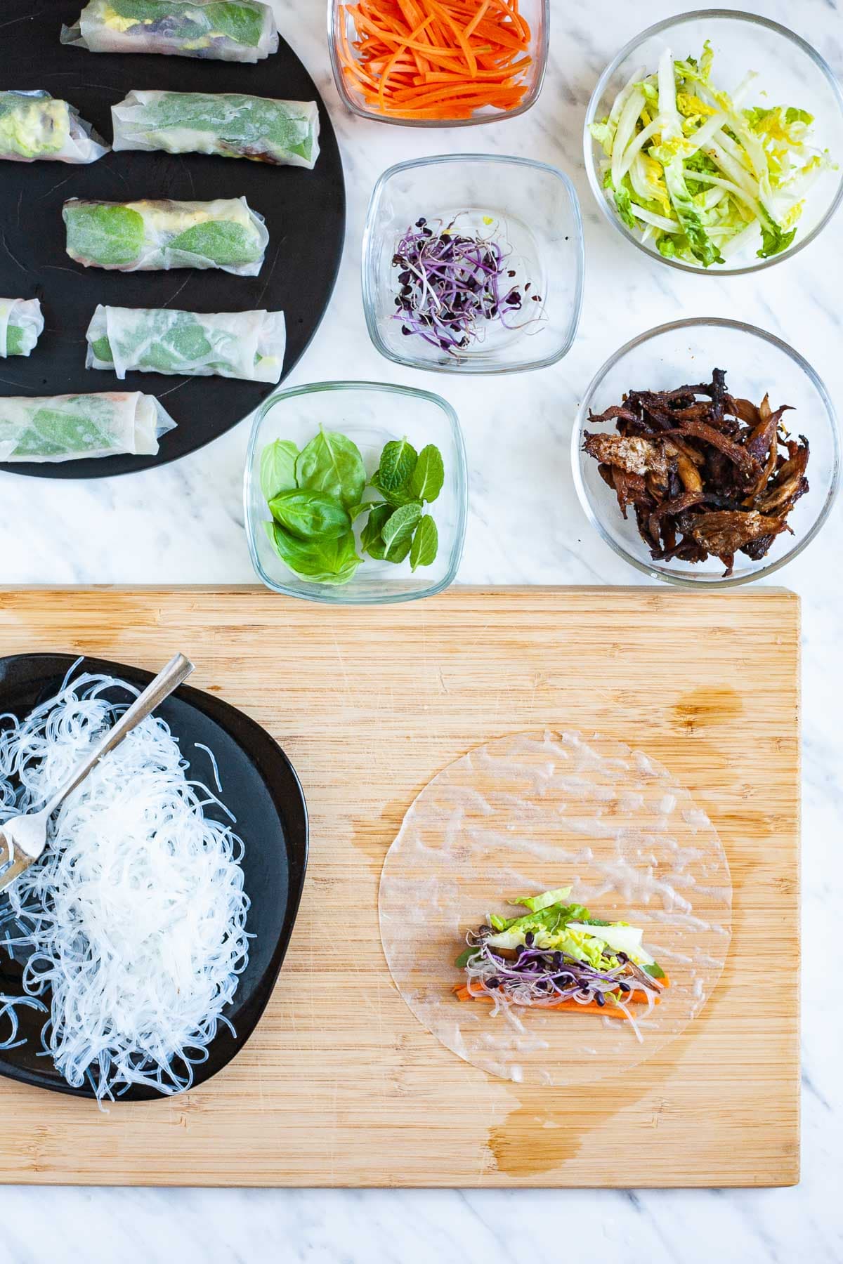 A wooden board with an opaque rice paper wrapper with a small about of filling. Leftover fillings are around it in small glass bowls. Glass noodles on a black plate. The finished summer rolls are in the corner.