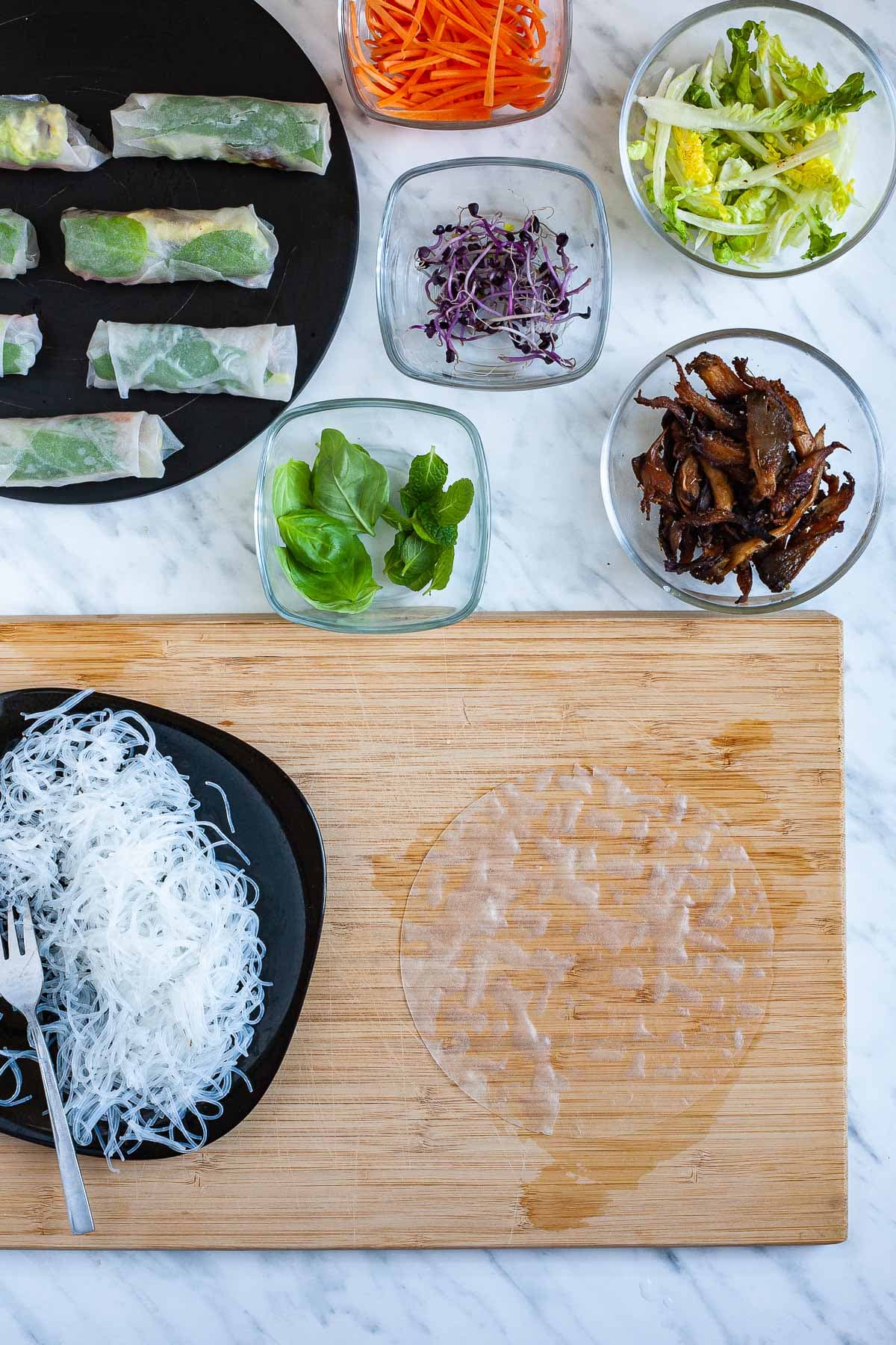 A wooden board with an opaque rice paper wrapper. Fillings are around it in small glass bowls. Glass noodles on a black plate. The finished summer rolls are in the corner.