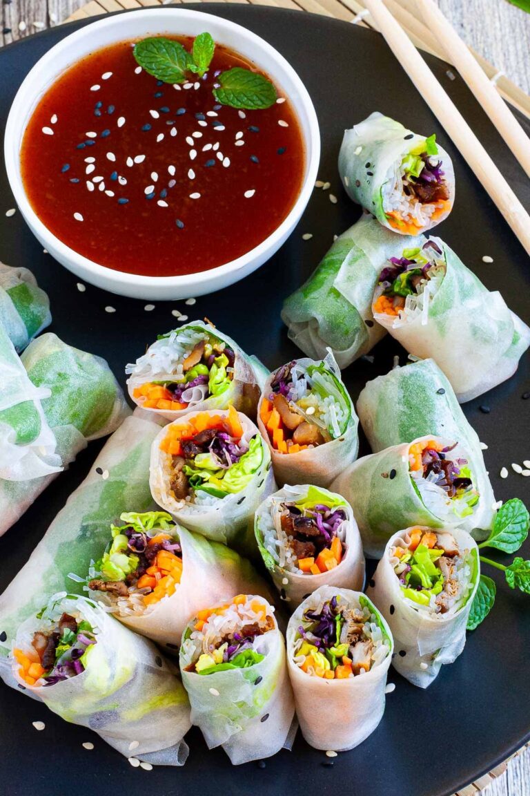 Opaque rice paper rolls filled with carrots, purple sprouts, thin glass. noodles and brown mushrooms are served on a large black plate. Several rolls are cut half so you can see the filling. Red sauce in a small white bowl is served next to it.