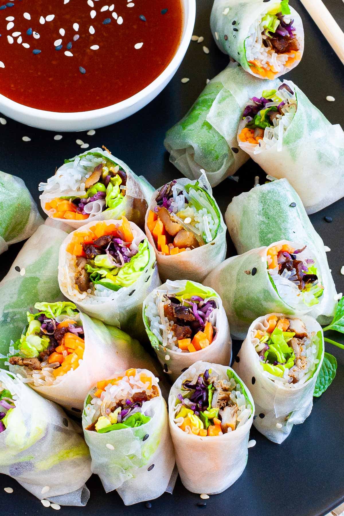 Opaque rice paper rolls filled with carrots, purple sprouts, thin glass. noodles and brown mushrooms are served on a large black plate. Several rolls are cut half so you can see the filling. Red sauce in a small white bowl is served next to it.