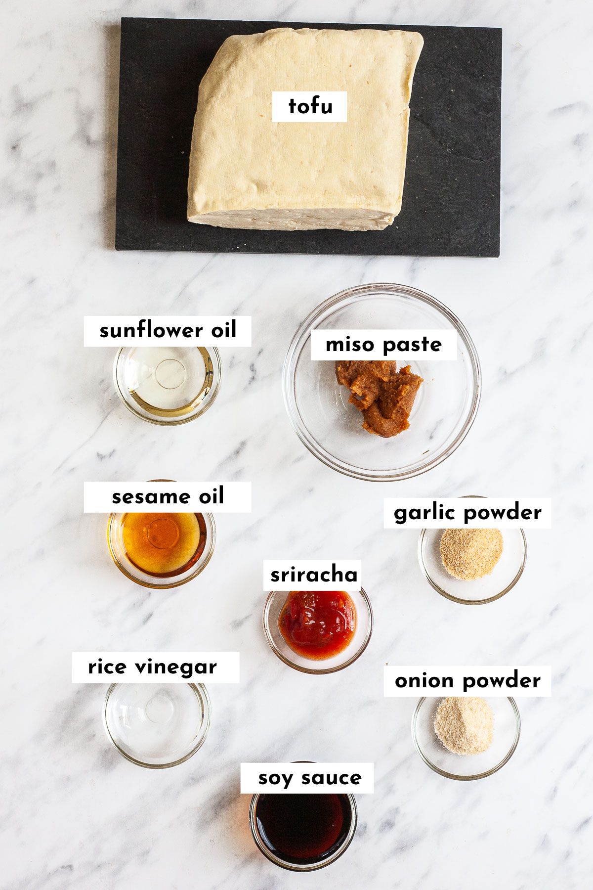Ingredients of miso tofu placed in glass bowls: miso paste, sriracha, sunflower oil, sesame oil, garlic powder, rice vinegar, onion powder, soy sauce and tofu on a black plate