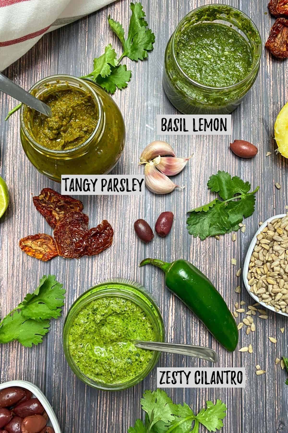 3 types of green pesto in small glass jars and different ingredients are around them like herbs, pepper, sundried tomatoes