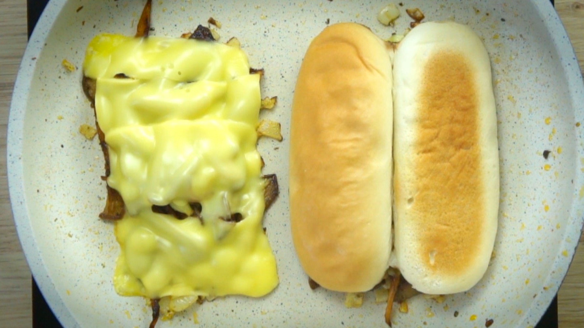 Shredded mushrooms separated into two parts covered with melted cheese and a hoagie roll in a white frying pan.