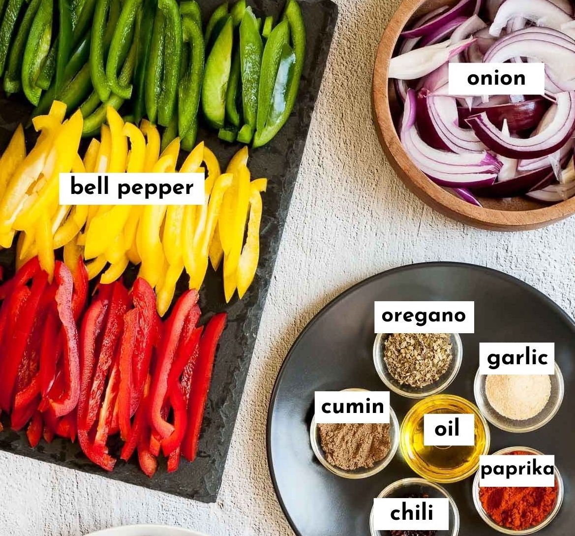The ingredients of fajita veggies in different plates. The spices in small glass bowls, the colorful bell peppers slices on a black board. Purple onion slices in a wooden bowl.