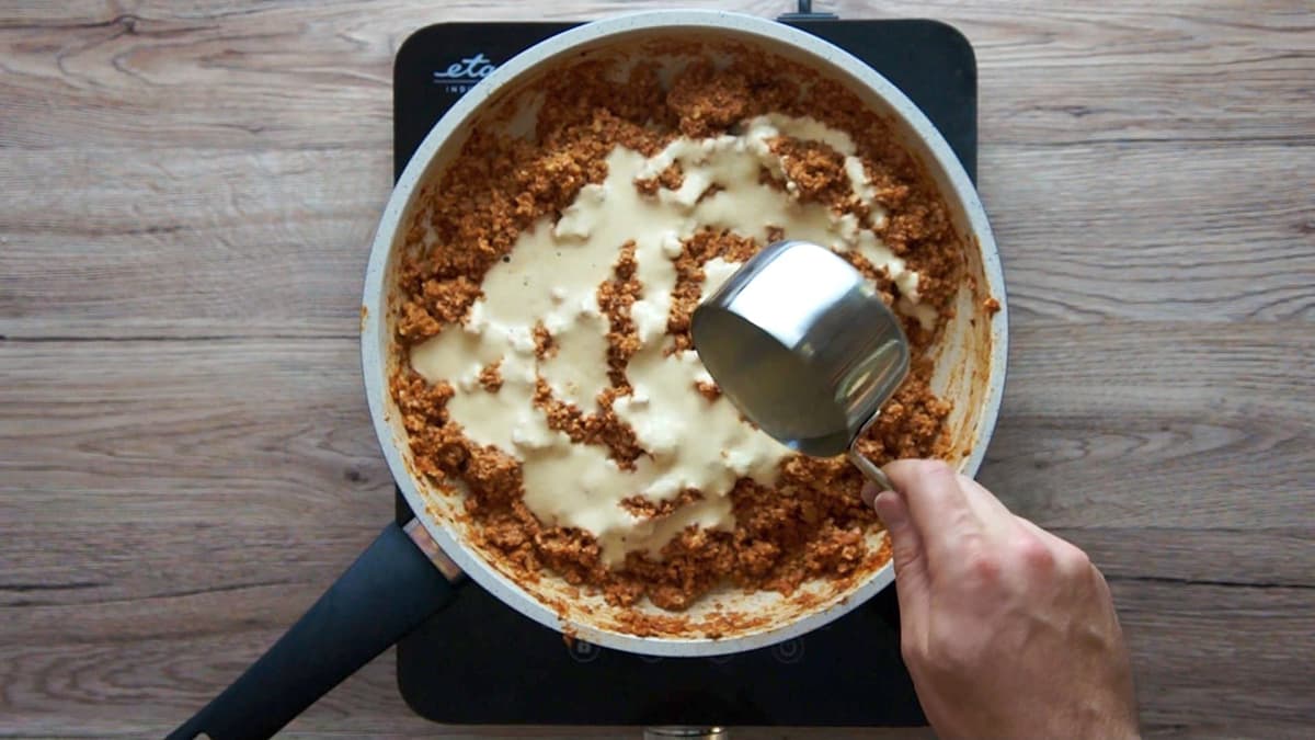 White frying pan with chopped cauliflower and walnuts in a red sauce. A hand is holding a cup and adding some white sauce. 
