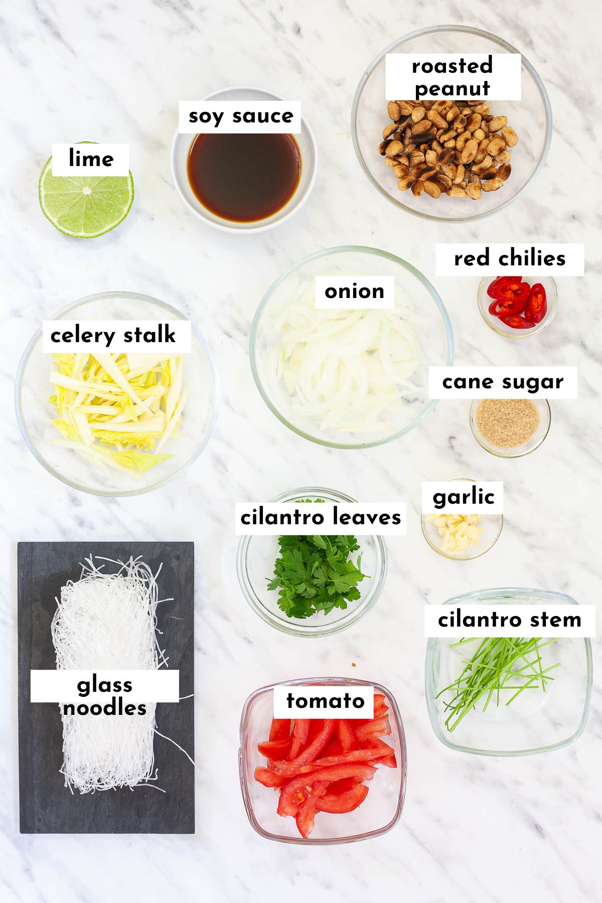Ingredients of yum woon sen in small glass bowls like peanuts, glass noodles, sliced tomato, cilantro leaves, stems, sliced celery, soy sauce, sliced onion, sliced chilies, minced garlic, sugar and half of lime, glass noodles