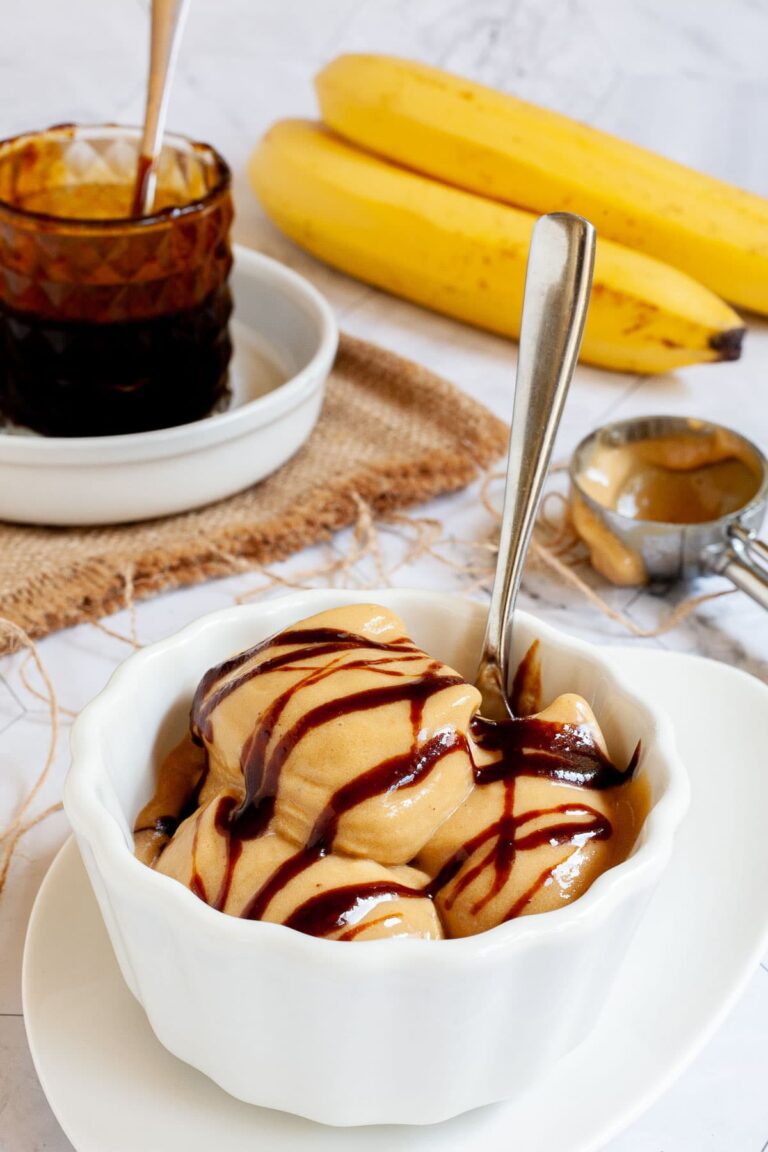 A white bowl of caramel colored ice cream with dark brown caramel drizzle on top. A glass jar of caramel sauce and 2 bananas are in the background.