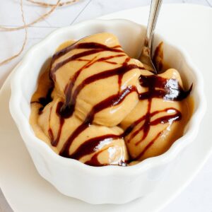 A white bowl of caramel colored ice cream with dark brown caramel drizzle on top.