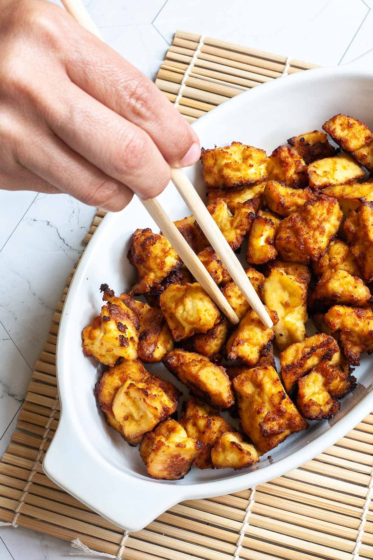 White bowl with yellow-brown tofu bites. A hand is holding chopsticks and taking one.