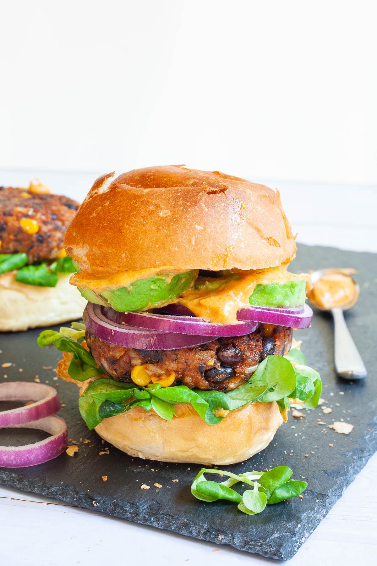 A burger bun with yellow sauce and green leaves is topped with a thick brown burger patty with visible sweet corn and black bean pieces, then red onion and avocado slices drizzled with yellow sauce again. It is covered with a top bun.