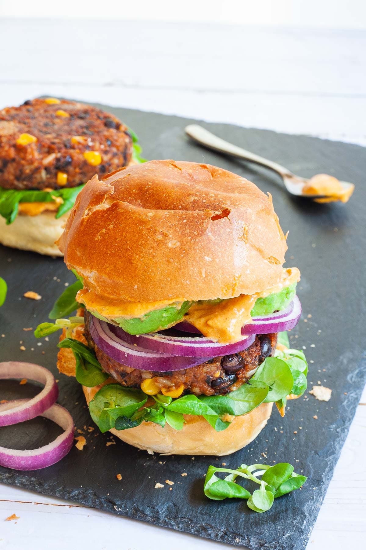 A burger bun with yellow sauce and green leaves is topped with a thick brown burger patty with visible sweet corn and black bean pieces, then red onion and avocado slices drizzled with yellow sauce again. It is covered with a top bun.