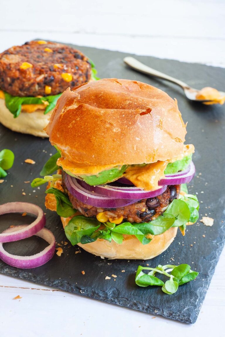 A burger bun with yellow sauce and green leaves is topped with a thick brown burger patty with visible sweet corn and black bean pieces, then red onion and avocado slices drizzled with yellow sauce again. It is covered with a top bun.