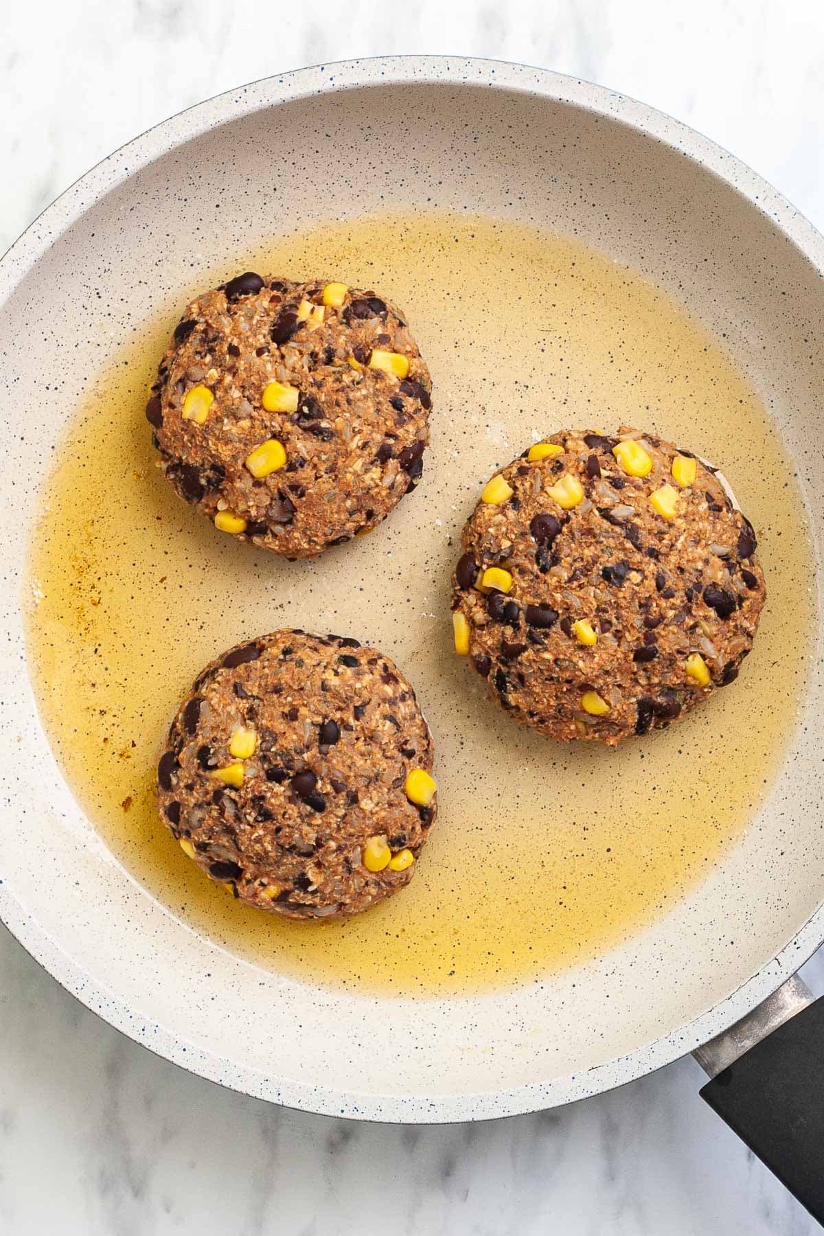 3 palm-sized brown burger patties are in a frying pan with oil. Sweet corn and black beans are visible inside.
