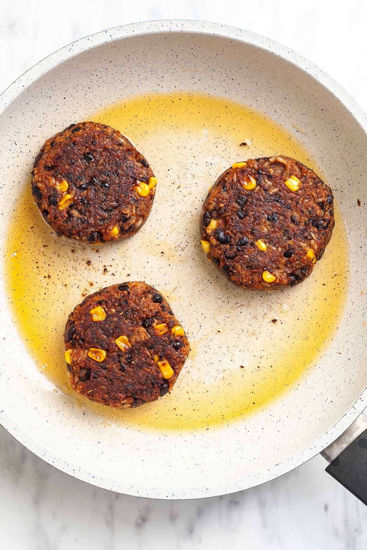 3 palm-sized dark brown crispy burger patties are in a frying pan with oil after flipping. Sweet corn and black beans are visible inside.