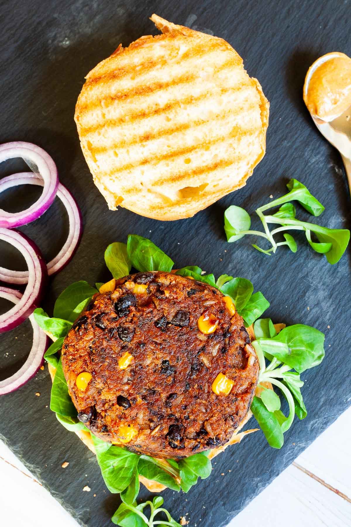 A burger bun with yellow sauce and green leaves is topped with a thick brown burger patty with visible sweet corn and black bean pieces. The top bun is next to it.
