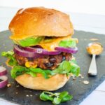 A burger bun with yellow sauce and green leaves is topped with a thick brown burger patty with visible sweet corn and black bean pieces, then red onion and avocado slices drizzled with yellow sauce again. It is covered with a top bun.