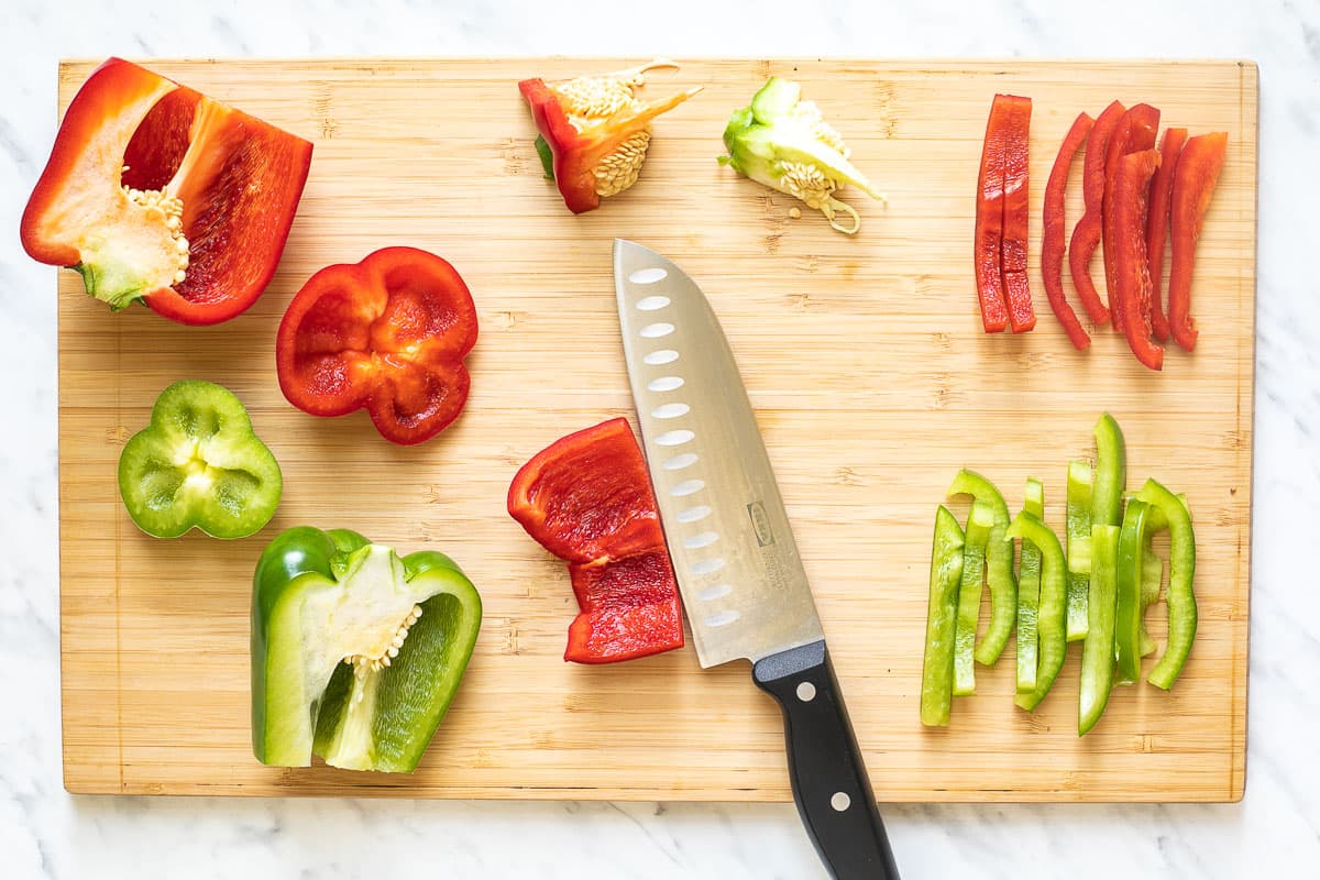 A wooden cutting board with a knife and green and red bell peppers cut into half and slices.
