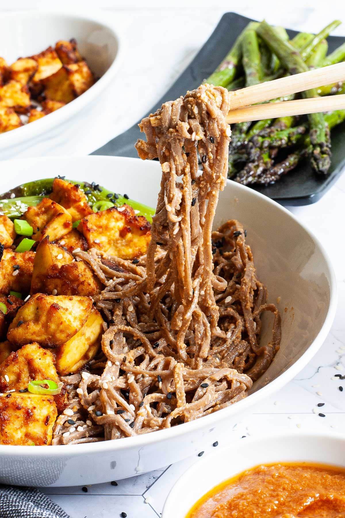 A white plate with brown noodles, yellowish crispy tofu bites. A hand is holding chopsticks and lifting some noodles up. Asparagus is in the background in a black plate. 
