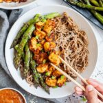 Large white bowl with brown soba noodles, orange crispy tofu bites, roasted asparagus and black and white sesame seeds. More asparagus and tofu bites are on the side. A hand is holding chopsticks and taking one tofu.