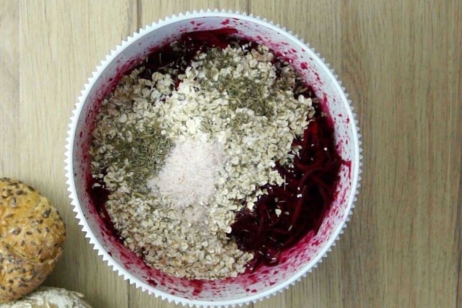 White mixing bowl with purple mixture for burger patties topped with oats, green dried herbs and salt.
