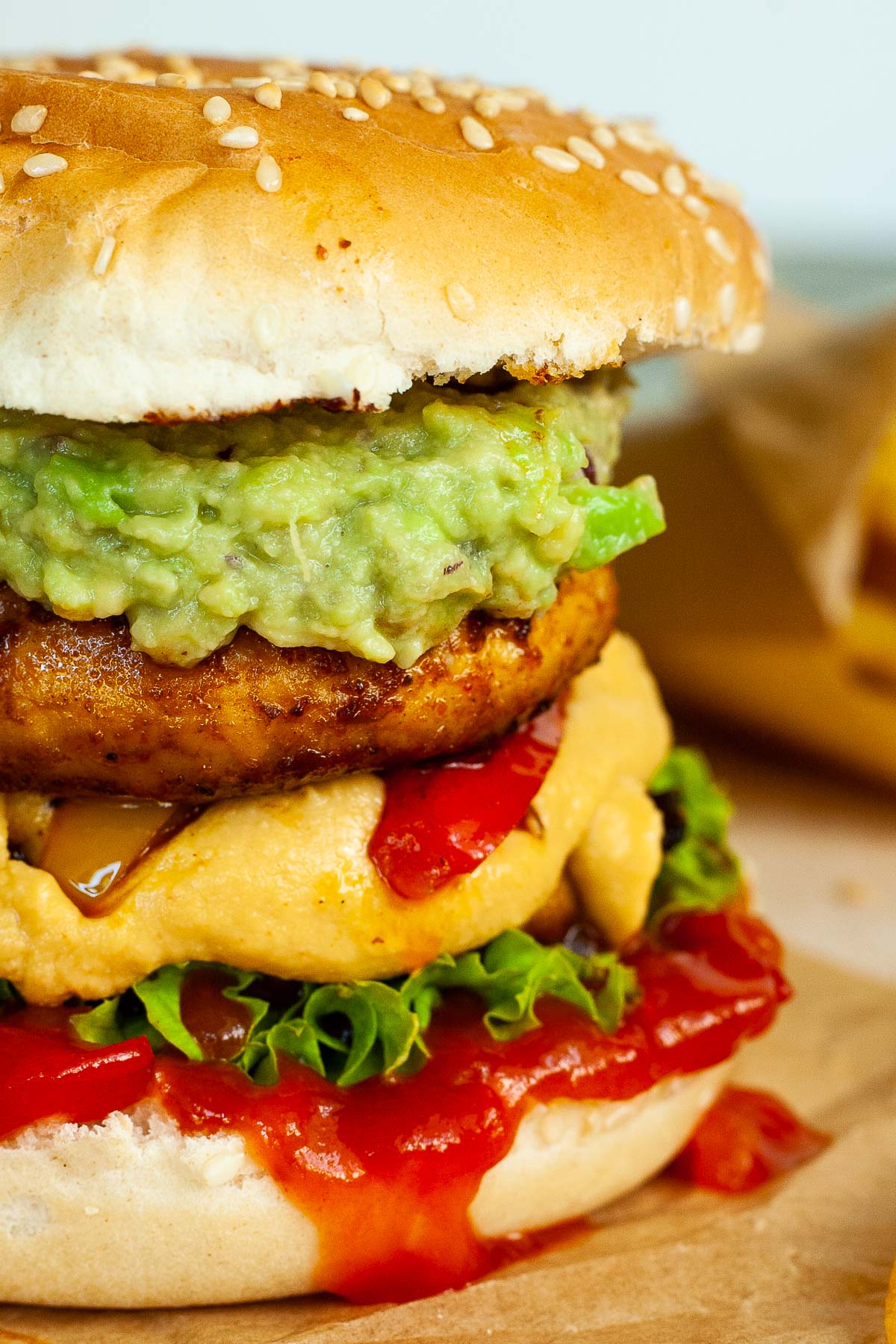 A burger with ketchup, lettuce, yellow cheese sauce, bell peppers, guacamole and grilled mushroom cap on brown paper. French fries are placed next to it.
