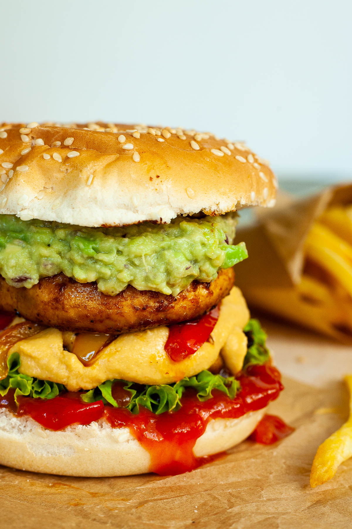 A burger with ketchup, lettuce, yellow cheese sauce, bell peppers, guacamole and grilled mushroom cap on brown paper. French fries are placed next to it.