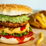A burger with ketchup, lettuce, yellow cheese sauce, bell peppers, guacamole and grilled mushroom cap on brown paper. French fries are placed next to it.