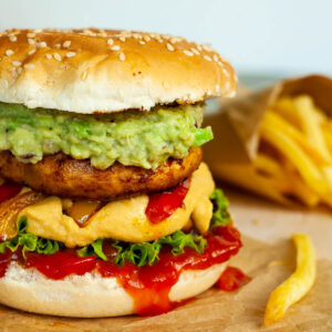 A burger with ketchup, lettuce, yellow cheese sauce, bell peppers, guacamole and grilled mushroom cap on brown paper. French fries are placed next to it.