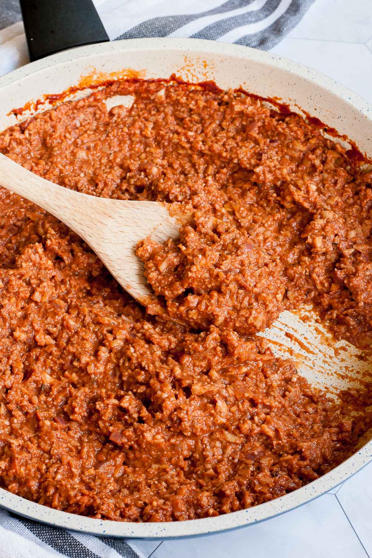 White frying pan with red thick sauce with fake ground meat pieces. A wooden spatula is taking some from the middle.