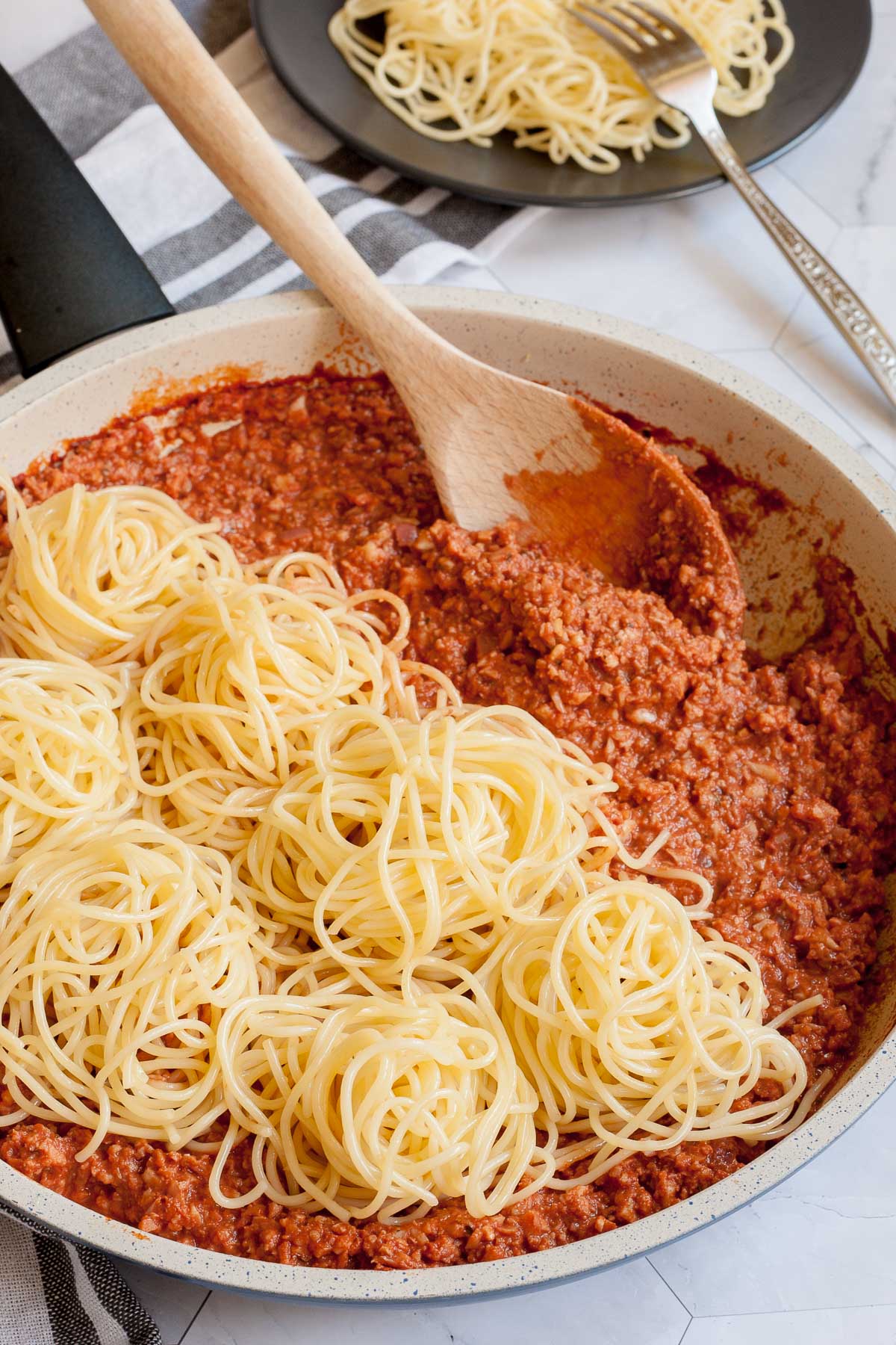 White frying pan with red thick sauce with fake ground meat pieces topped with small heaps of spaghetti. A wooden spatula is placed inside.