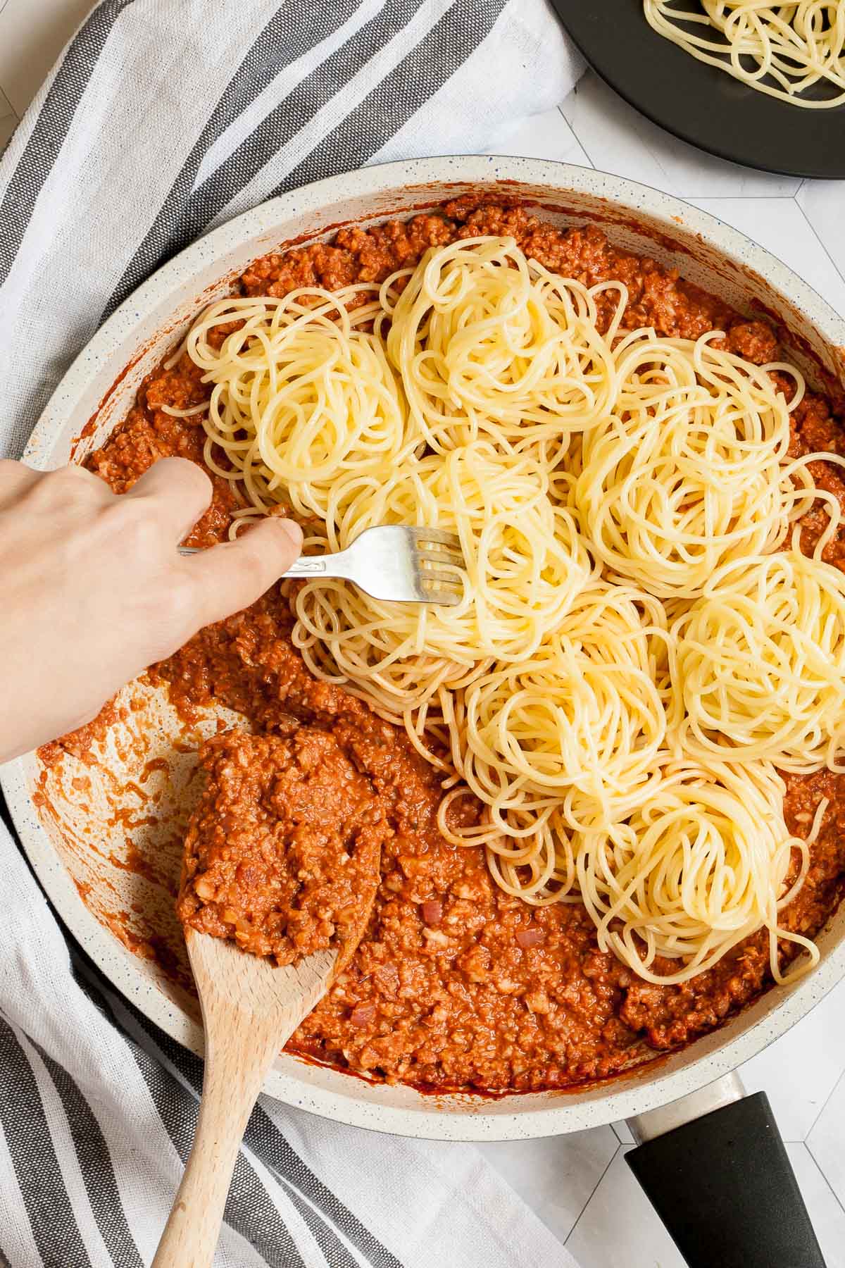 White frying pan with red thick sauce with fake ground meat pieces topped with small heaps of spaghetti. A wooden spatula is placed inside. A hand is holding a fork and pointing in the pasta.