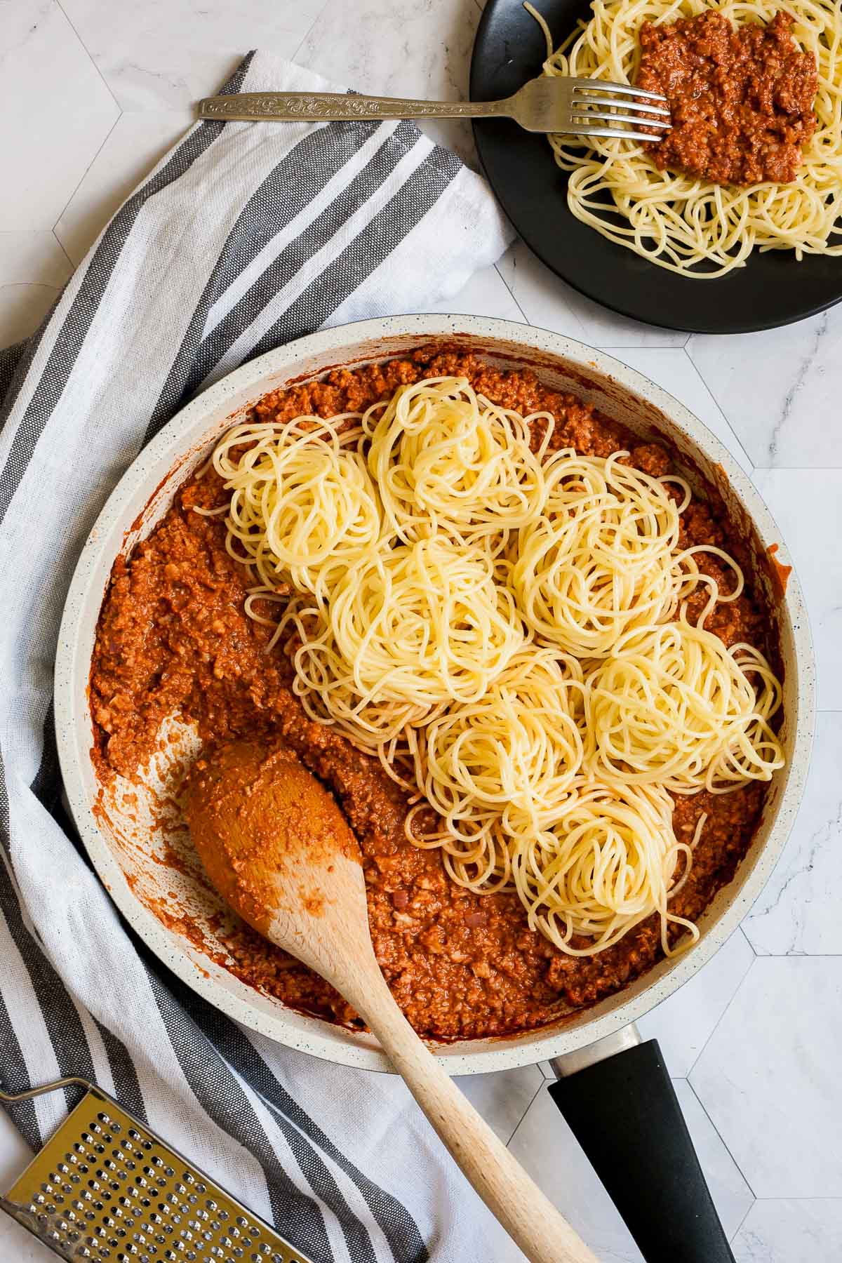 White frying pan with red thick sauce with fake ground meat pieces topped with small heaps of spaghetti. A wooden spatula is placed inside. A small black plate with a serving serving is placed next to it. 