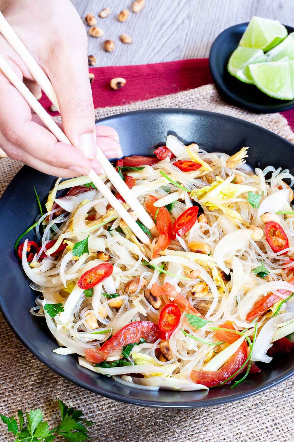 Black plate on a red mate with thin glass noodles, sliced tomatoes, roasted peanuts, sliced onion, sliced chili peppers, chopped green herbs. A hand is holding chopsticks to start eating.