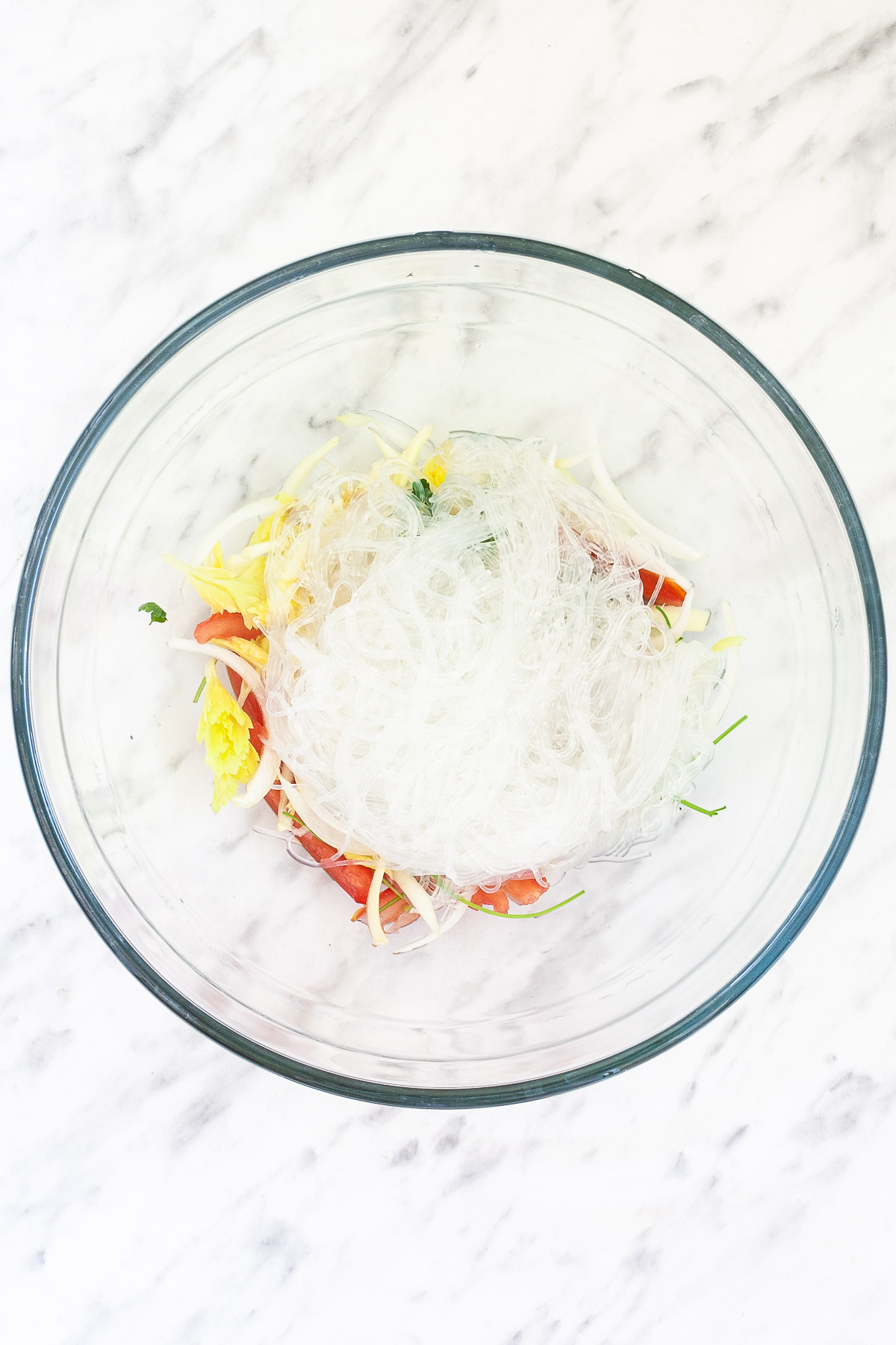 Glass bowl with tomato slices, cilantro, celery, and sliced onion topped with a heap of thin glass noodles. 