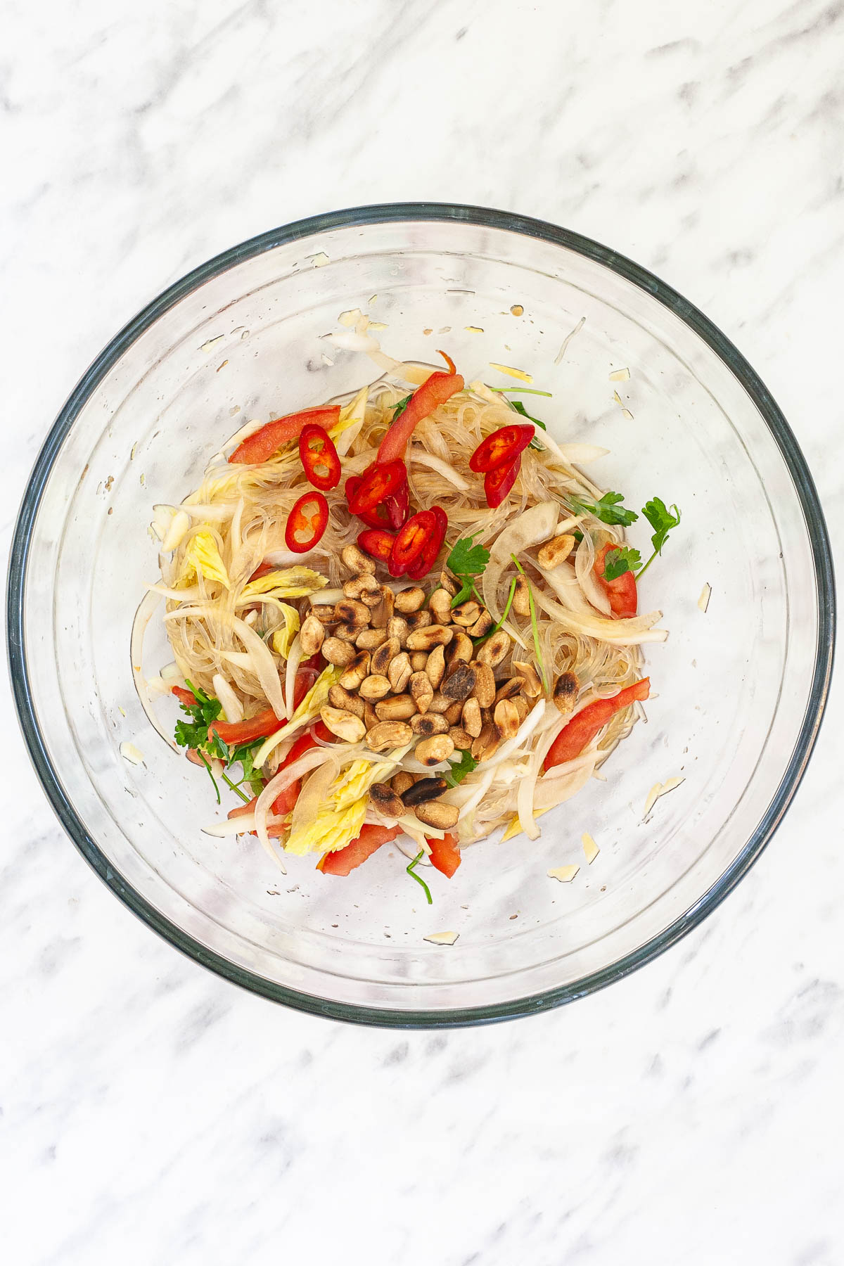 Glass bowl with tomato slices, cilantro, celery, sliced onion and thin glass noodles slightly brown from a thin sauce topped with roasted peanuts and chili pepper slices. 