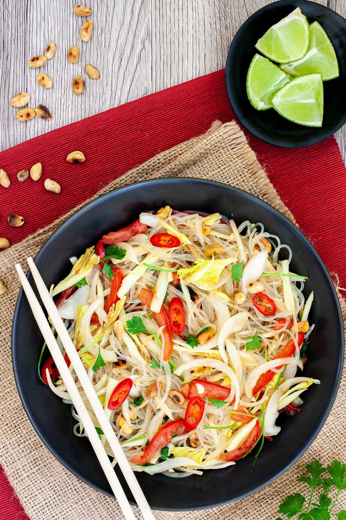 Black plate on a red mate with thin glass noodles, sliced tomatoes, roasted peanuts, sliced onion, sliced chili peppers, chopped green herbs. Chopsticks are placed on the side.