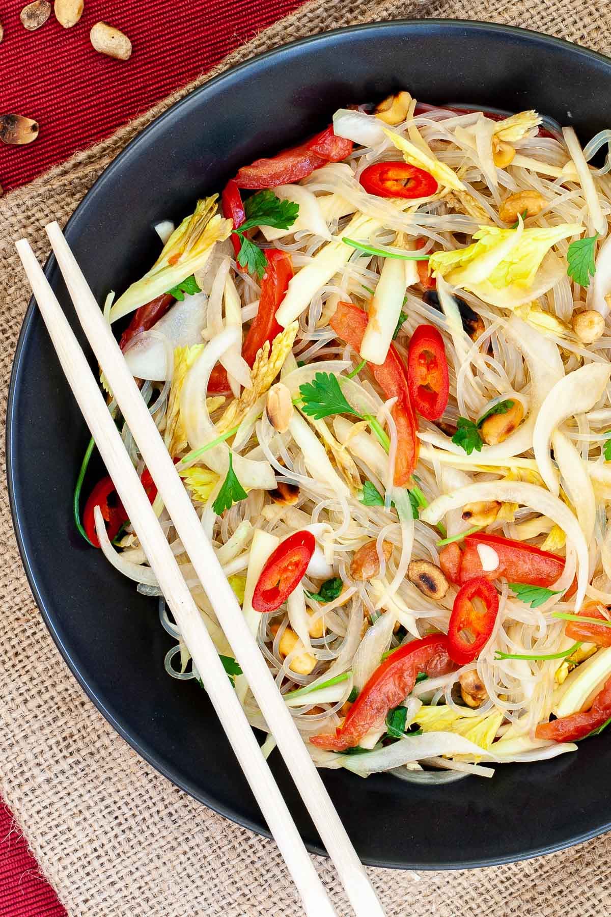 Black plate on a red mate with thin glass noodles, sliced tomatoes, roasted peanuts, sliced onion, sliced chili peppers, chopped green herbs. Chopsticks are placed on the side. 