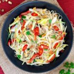 Black plate on a red mate with thin glass noodles, sliced tomatoes, roasted peanuts, sliced onion, sliced chili peppers, chopped green herbs.