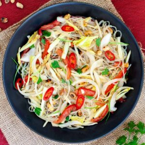 Black plate on a red mate with thin glass noodles, sliced tomatoes, roasted peanuts, sliced onion, sliced chili peppers, chopped green herbs.
