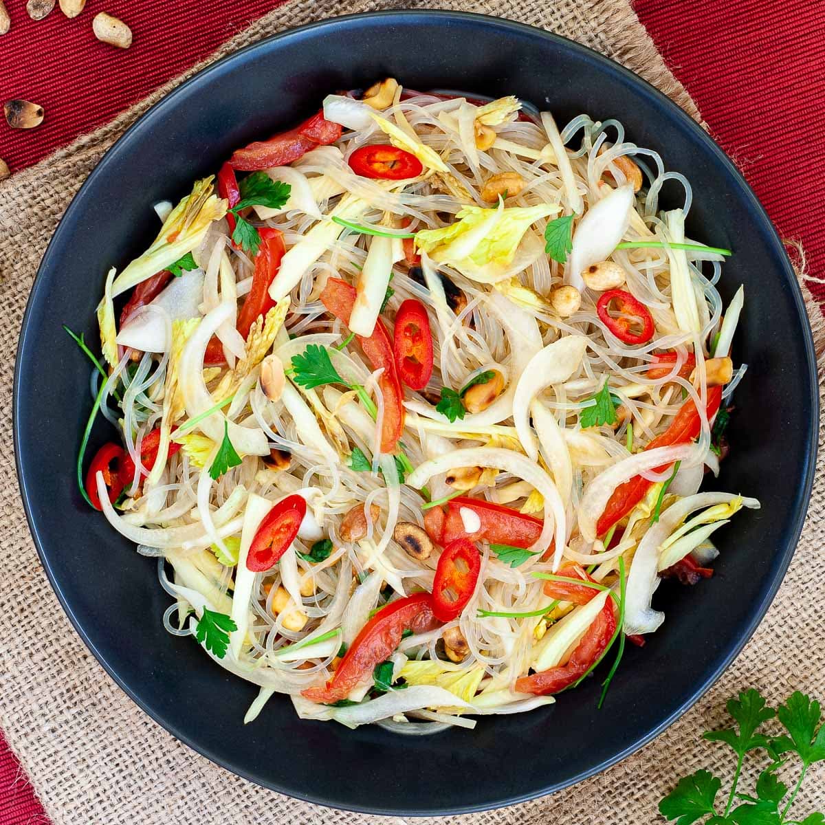 Black plate on a red mate with thin glass noodles, sliced tomatoes, roasted peanuts, sliced onion, sliced chili peppers, chopped green herbs.