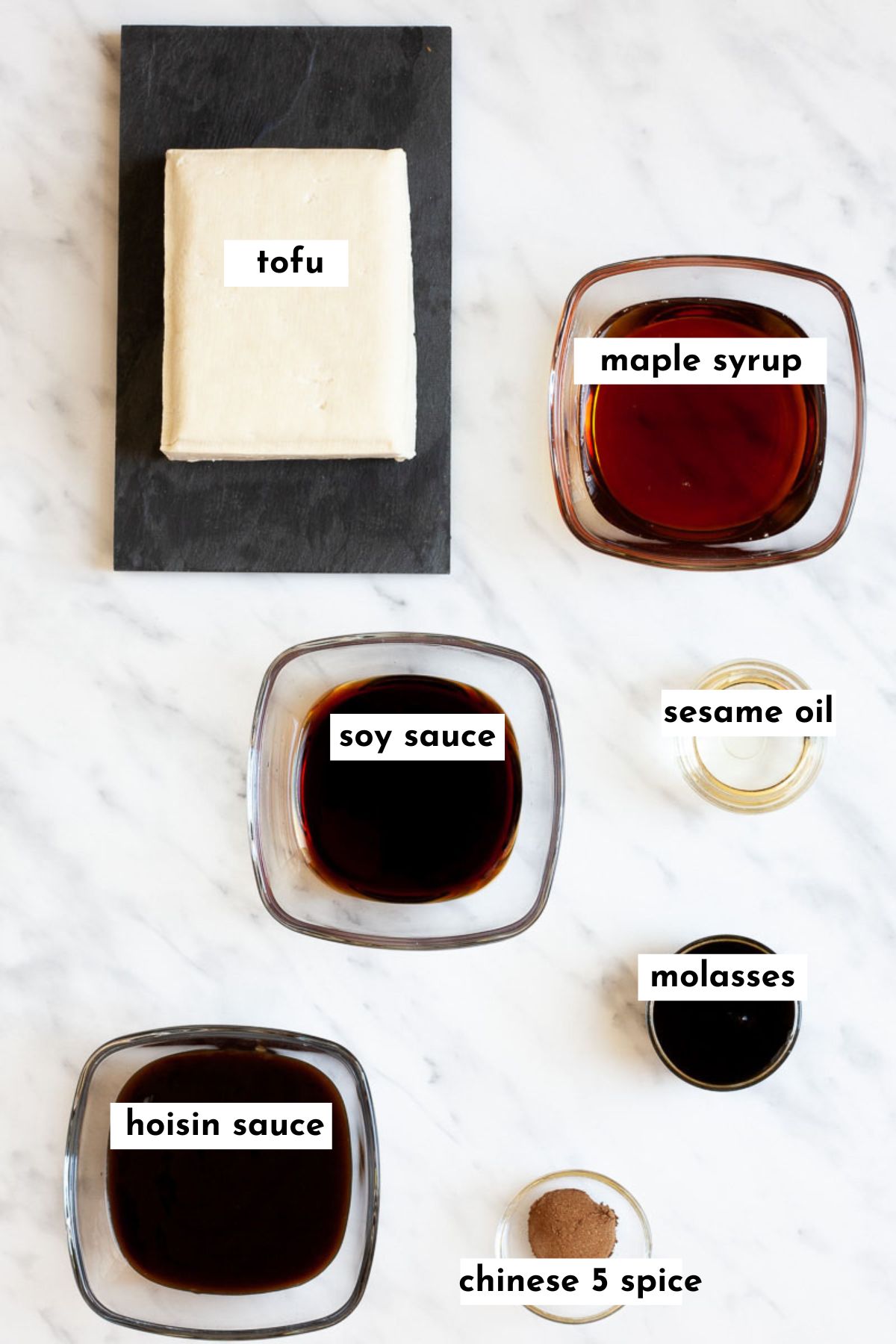 The ingredients of the tofu burger is placed in glass bowls like maple syrup, soy sauce, molasses, hoisin sauce, spice and oil. A brick of tofu is placed on a black plate. 